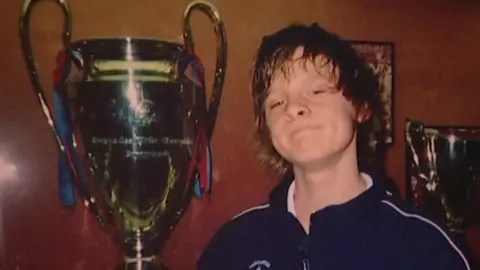 A large trophy with two handles, resembling the UEFA Champions League cup, is prominently displayed in the centre. The trophy has ribbons in blue and red attached to its handles. Joe stands to the right, he is wearing a dark sports jacket with a white zipper and a crest on the chest.