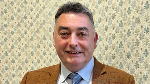 Local Democracy Reporting Service Councillor Danny Brookes, who has short, grey hair, wearing an orange, checked blazer, a white shirt, and a blue and white patterned tie. He is smiling at the camera and is stood in front of a wall with a blue and yellow leaf pattern on.