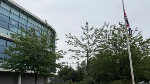 Mark Denten/BBC A modern office building with a glass front made from rows of windows has small trees in front and, to the side, a white flag pole with a union jack flag at half mast.