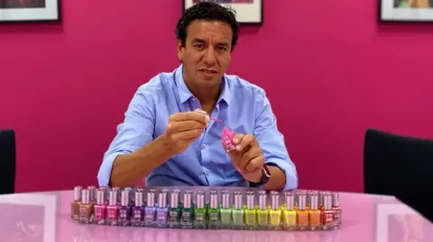 BBC Man with dark hair and brown eyes, wearing a mid-blue button-up shirt, sits at a table on which there is an array of nail vanishes in different colours including pink, purple, lilac, green, yellow and orange polishes. 