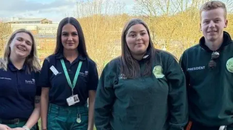 Three women and one man dressed in green paramedic outfits, standing in front of a fence above a road.