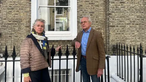 Two neighbouring residents standing beside a black iron railing outside a brick townhouse, with a window visible behind them.