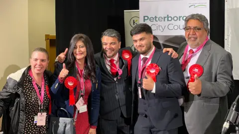 Ben Schofield/BBC Five people, wearing Labour rosettes pose for a picture. Four of them are giving a thumbs up sign. In the background are banners advertising Peterborough City Council. 