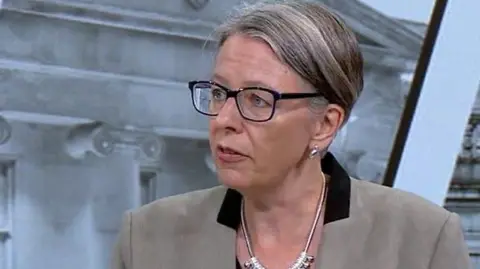 Geraldine Coggins, wearing glasses and a brown jacket with a black collar