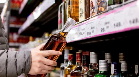 Getty Images A person holds a bottle of amber‑coloured alcohol while browsing a supermarket shelf filled with various spirits, with price labels displayed along the rack.