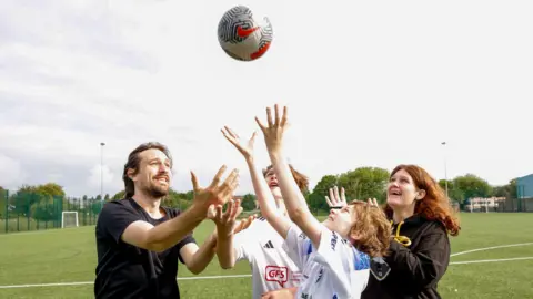 A man, two boys and a woman stand on a football pitch with their arms reaching up to a ball, which is in the air. The man and woman are in black, while the boys wear white football shirts. 