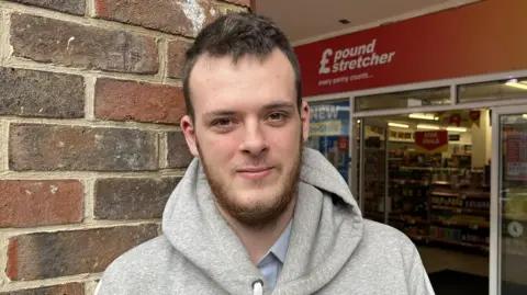 Phil Harrison/BBC A man with a brown beard and short brown hair. He is wearing a grey hoodie and standing in front of a Pound Stretcher.