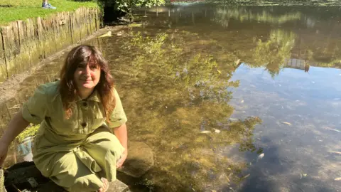 Owen Sennitt/BBC A woman wearing a light green jumpsuit kneeling down by the side of the water. She has wavy brown hair and is smiling while looking up to the camera. She is kneeling next to the river.