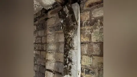 Keele University The black fabric remains of blackout curtains, that are nailed to a light wooden doorframe which is on a brick wall. The fabric is aged and in tatters, with white cobwebs all over it