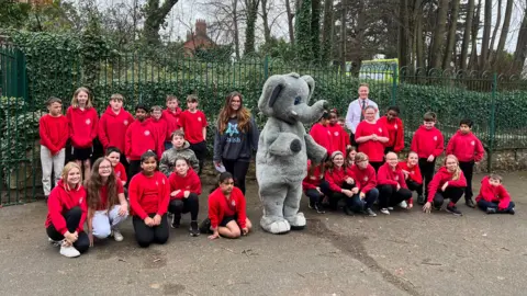 A group picture of students from St John's Primary School in Wellington, Somerset. There is an elephant mascot standing amongst the group of children, who are all in red uniforms. Naomi from 2Wish is next to the elephant mascot. She has long brunette hair and is wearing a navy blue hoodie with a star logo on for 2Wish. Harry Foster is stood in the group. He has fair hair and is wearing a shirt and tie. Most of them are looking at the camera. 