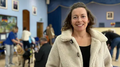 Elle is wearing a light-colored fleece coat stands in a hall with blue walls and framed pictures. In the background, several people interact with dogs near blue chairs and tables, with a stage visible at the far end.