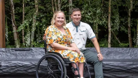 Chloe Docker Chloe Docker sitting in her wheelchair next to her husband. She has blonde hair and is wearing a yellow and orange floral dress. Her husband has his arm around her and is wearing a white polo shirt and khaki trousers.