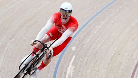 PA Media A man wearing a red cycling suit and white helmet is seen riding on an indoor cycling track, tilted at an angle. He has his mouth open, his face is obscured by a visor.