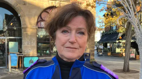 A woman with dark hair and wearing a blue top with two different shades of blue is looking at the camera.