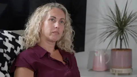 A woman with blonde curly hair and maroon dress sits in a sofa with a spiky pot plant behind her