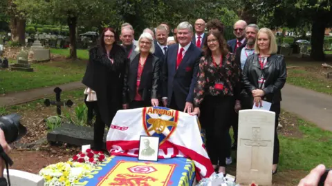 A group of men and women are standing in a cemetery next to a grave that is covered in an Arsenal flag with a picture of David Danskin