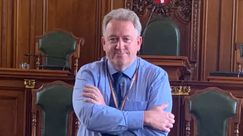 A man in a blue shirt and blue tie with his arms crossed smiling in a council chamber.