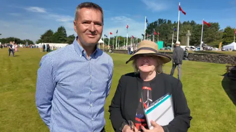 Ian Stratford & Georgina O'Sullivan stand side by side smiling, in front of a row of flagpoles, and blue skies. 