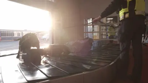 Isle of Man Government A black dog on a lead sniffs suitcases on an arrivals hall conveyor belt. A police officer wearing a hi-vis waistcoat holds the other end of the lead.