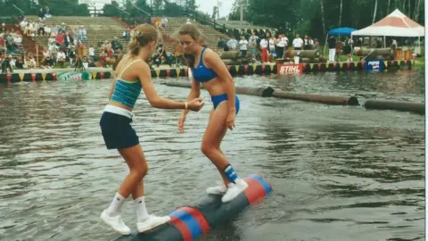 Samantha LaSalle Two women with their dirty blonde hair pulled back, one in a two-piece swimsuit and the other in shorts and a tank top, balance in socks and sneakers on a rolling log in water as stands of spectators look on