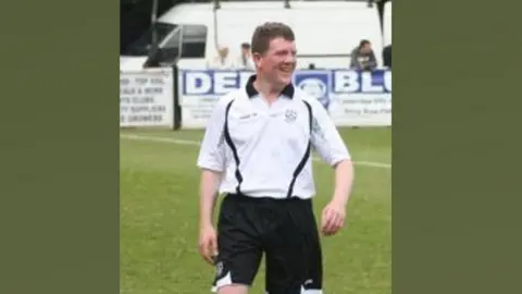 Pauline Muncey A blurry image of Neil Harvey in black and white football strip on the Milton Road pitch. He is looking towards the left and smiling. It was taken during a Cambridge City 'Legends' against BBC Radio Cambridgeshire at Milton Road in May 2010