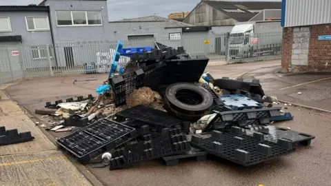 Tony Milson A pile of rubbish including tyres and plastic and wooden crates dumped on an industrial estate. There is a lorry in the background parked behind some metal railings.