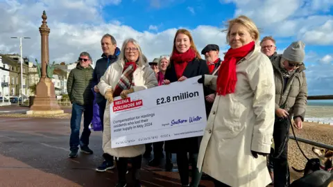 BBC/SARA SMITH Helena Dollimore stands on Hastings seafront with some of her constituents, holding a giant cheque for £2.8m from Southern Water.