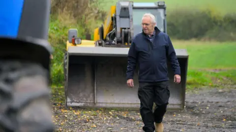 Rob Alderson walking along a field with a tractor behind him. It has a front-end loader attached to it. Rob has white hair and is wearing glasses. He is wearing dark trousers and a blue jacket over a blue checked shirt. Part of a tractor wheel is in the forefront of the picture.