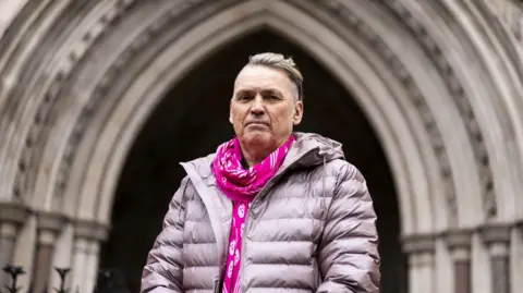 Dale Vince, the owner of green energy company Ecotricity, stands in front of the stone archway which is the main entrance to the High Court in London, wearing a light-coloured thick coat and a vibrant, pink-coloured scarf.