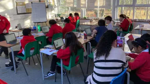 BBC Children are sitting in a classroom. They are wearing red jumpers and are working on computers.