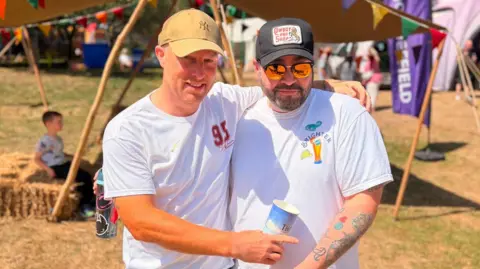 Two men in white t-shirts and caps. One points at the tattoo on Hefty's wrist.