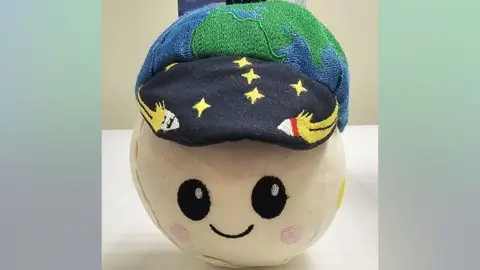 NASA A small, round soft toy sits on a white tabletop, about the size of a large grapefruit. Its fabric face is pale cream with big black embroidered eyes, rosy pink cheeks and a simple smiling mouth, giving it a cute cartoon look. On top, it wears a dark blue cap sprinkled with embroidered yellow stars and tiny white rockets. Above the cap is a padded green‑and‑blue globe, like a miniature Earth, with a short dark loop for hanging. The toy’s overall impression is friendly and playful, like a cheerful mascot for a children’s space adventure.