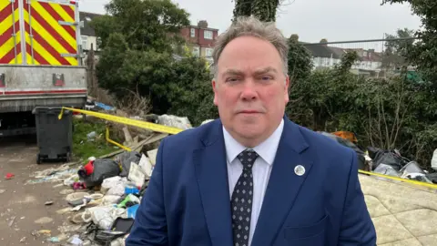 A large-built man in a blue suit with greying hair, stares into the camera. Behind him is discarded rubbish with yellow tape on it - and a rubbish truck to the left.