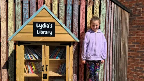 Lydia is in a pink hoodie standing in front of a rainbow-coloured wooden fence with 'Lydia's Library' next to her. It is a small wooden hut with two glass doors filled with books.