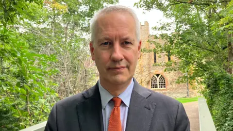 A man in a grey jacket and orange tie standing between trees in front of an old building