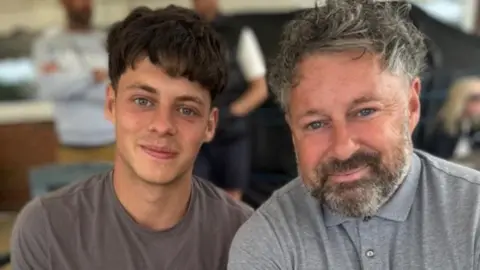 A man with curly hair and a grey flecked beard is on the right hand side of the picture, smiling. Alongside him is a younger man with dark hair, also smiling