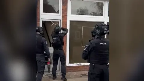 West Mercia Police Three police officers in black tactical equipment are stood outside a brick building with large white window frames. The officer closest to the building uses some equipment to break glass around the door.