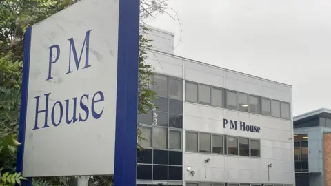Simon Thake/BBC A large office building with the name “PM House” displayed prominently on its exterior. The building has a modern, rectangular design with multiple rows of windows and light grey cladding. In the foreground, a tall sign also reads “PM House” in blue lettering on a white background, framed by a dark blue border. Several cars are parked in front of the building, suggesting a business or office complex. 