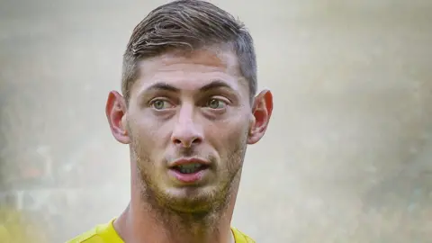 Emiliano Sala has short brown hair and is wearing a yellow Nantes Football Club top. Crowds of spectators at a football ground are blurred out behind him.