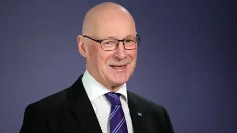 A bald man with glasses, wearing a dark suit, white shirt and purple stripy tie, looks at the camera in a medium close-up shot in front of a purple background 