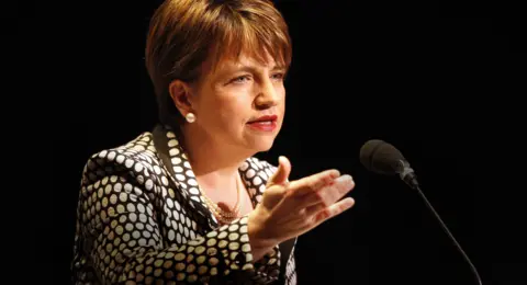 PA A woman with short dark hair and a dotted top speaks into a microphone