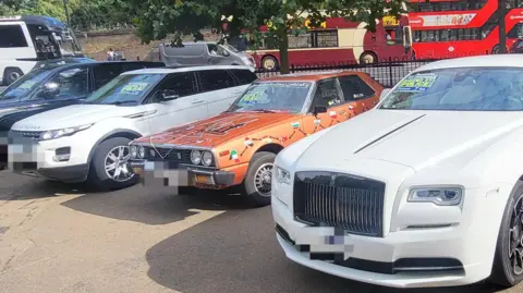 Met Police A row of seized cars with a white Rolls Royce closest on the right hand side of the picture, next to it an orange Nissan Datsun Skyline, then a white Range Rover. In the background are two buses and a silver van driving along a road.