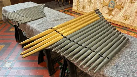 Lance Foy Organs Two rows of metal pipes are laid on a table. They resemble large pan pipes, with slits cut out a third of the way up.