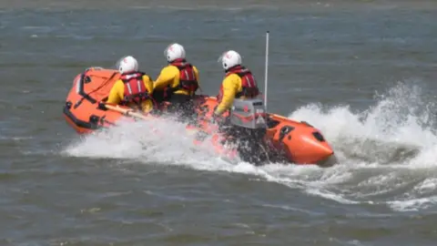 An orange lifeboat belonging to the RNLI showing three RNLI crew members on the sea, on the way to an incident. 
