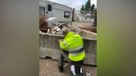 Environment Agency An environment agency worker, wearing a yellow hi-vis jacket, is knelt in front of a long concrete black, with his arms raised to it. Behind the knee-high block, there is a white caravan surrounded by waste including shards of wood, papers and white goods. 