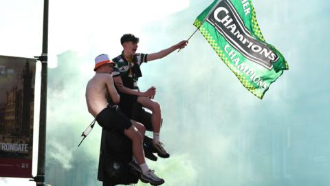 Two men sitting on a traffic light waving a flag reading 'premier champions'