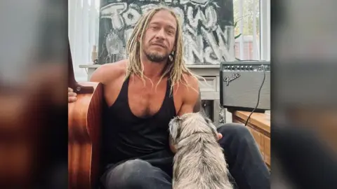 A man with long hair and a beard.  he strokes a dog