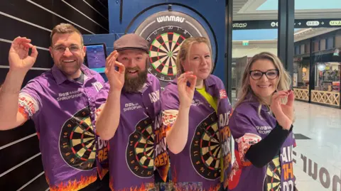 BBC Four people are wearing a mainly purple short-sleeved shirt that has BBC Radio Shropshire and an image of a dartboard on it. All of them have their right hand pointing to the camera as if they were holding a dart they were about to throw. A dartboard is behind them.
