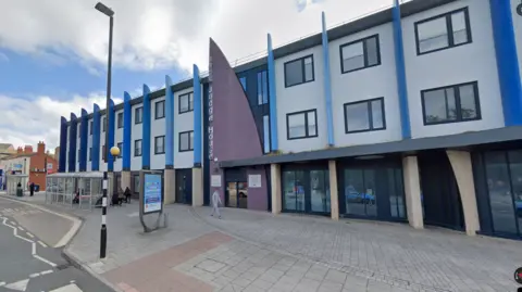 Google The Black Country Coroner's Court. It is a modern looking building. There is a bus stop nearby and a person walking in the street. 