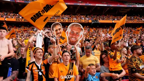 PA Tigers fans in the stands at Wembley holding flags and a large cardboard cut out of Oli McBurnie's face